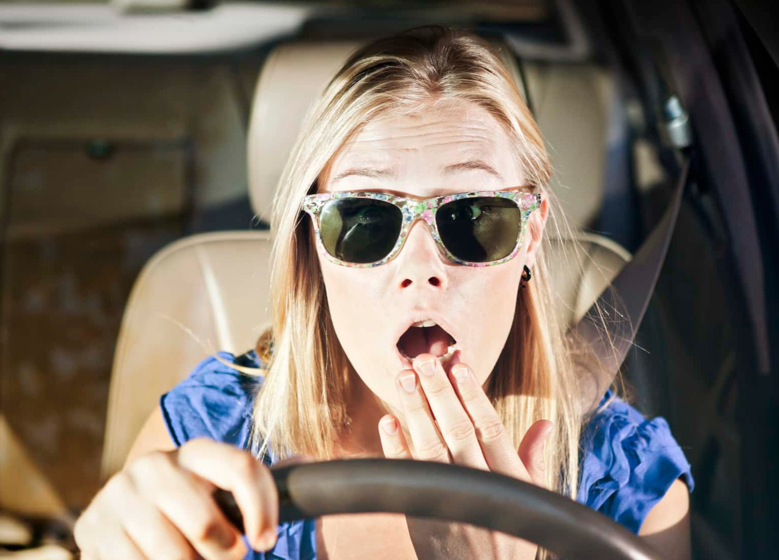 Horrified blonde drive with hand to mouth, looks through windscreen.