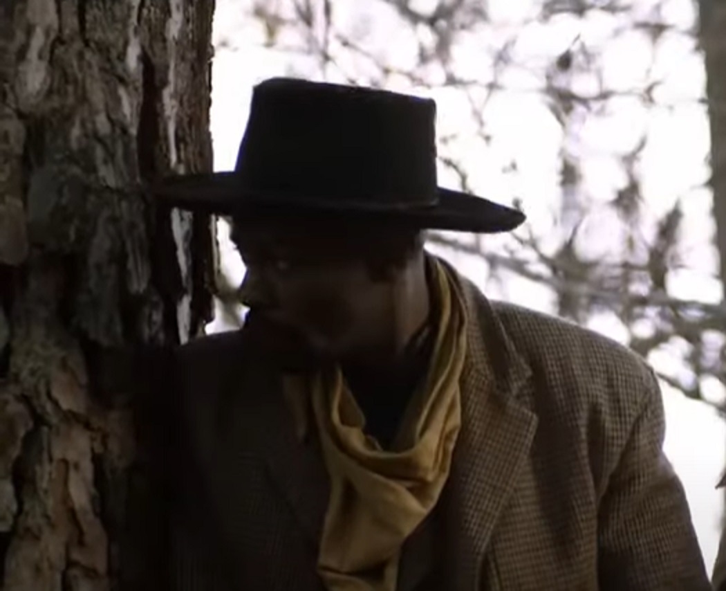 Screenshot:Bass Reeves wearing brown jacket and hat standing outside - from H.e.l.l on the Border (2019)