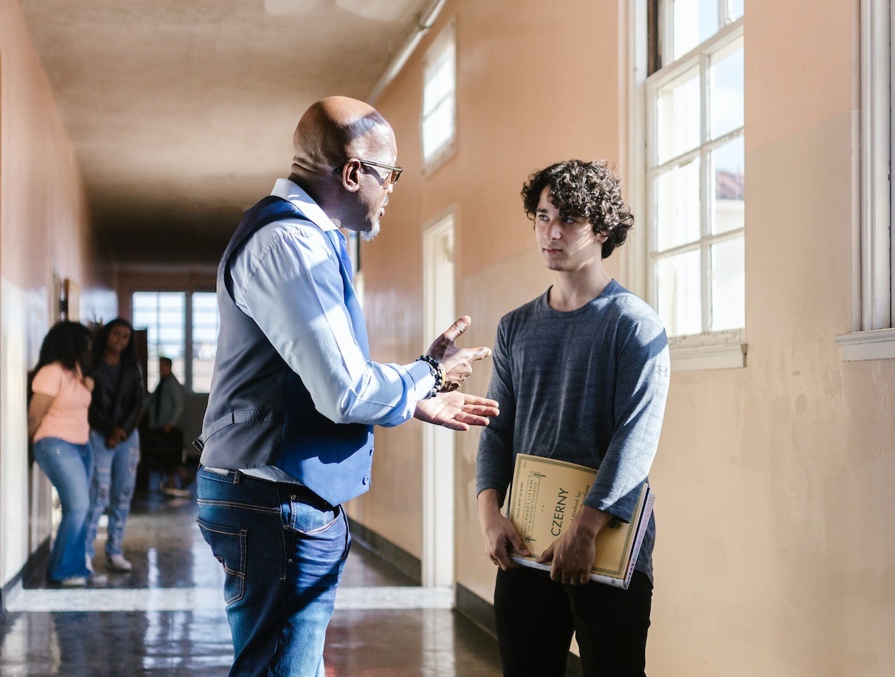 Teacher in blue shirt is talking with student in school hallway.