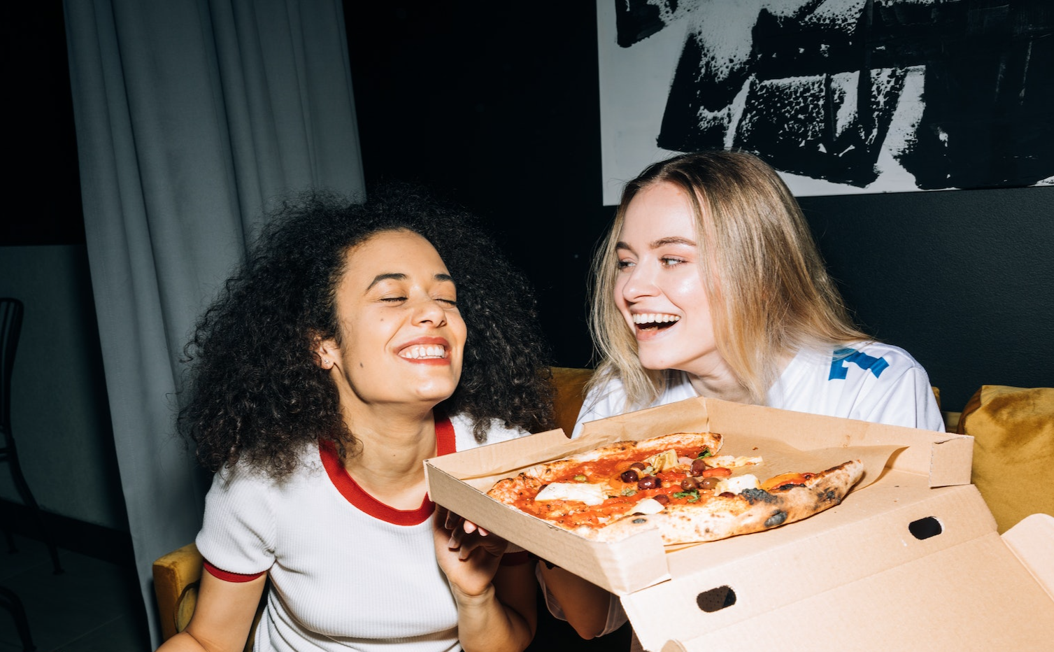 two young women having fun and eating pizza in sitting on a couch