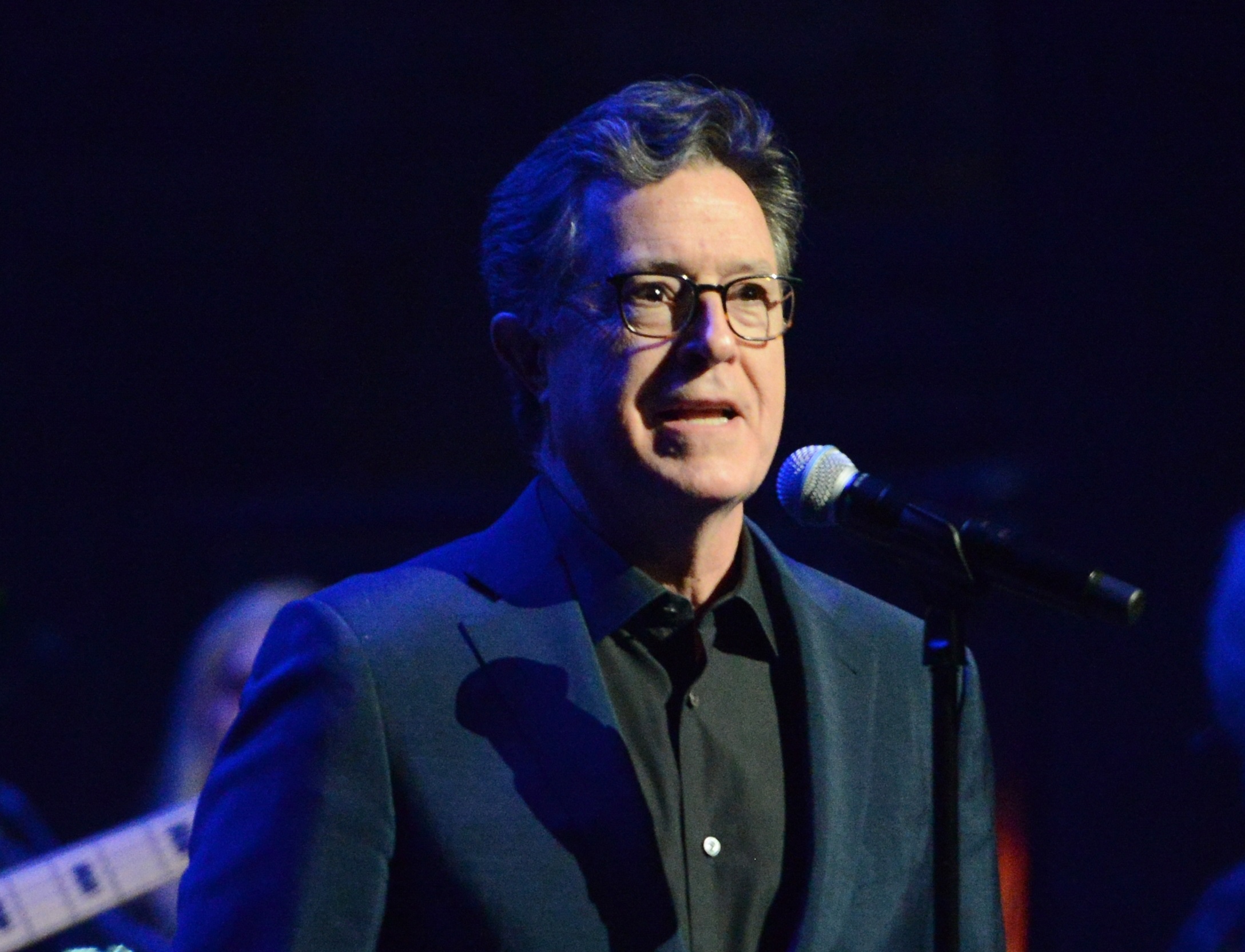 Stephen Colbert wearing black suits is looking at side and talking - 2023
