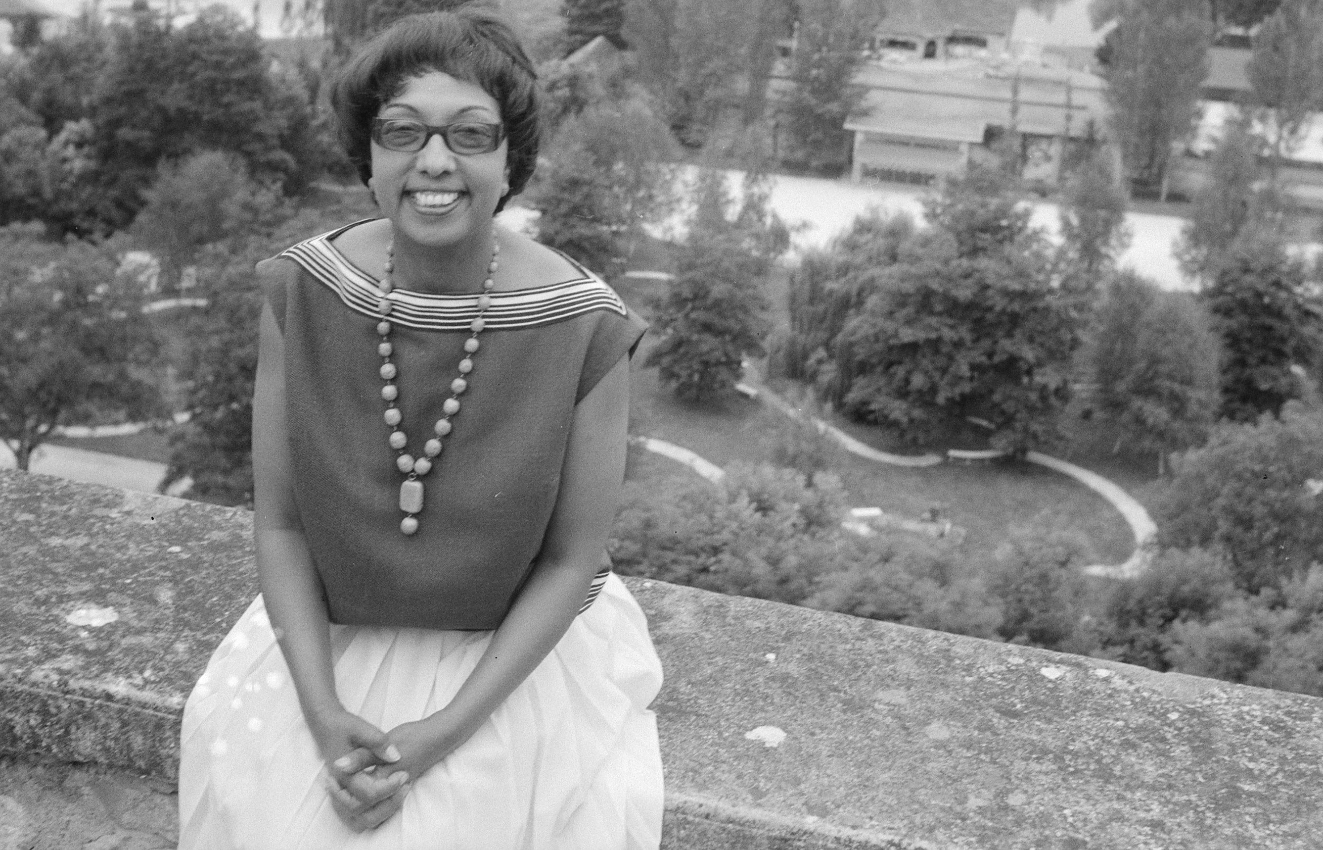 Grayscale photo of elder Josephine Baker sitting on a wall smiling and facing the camera