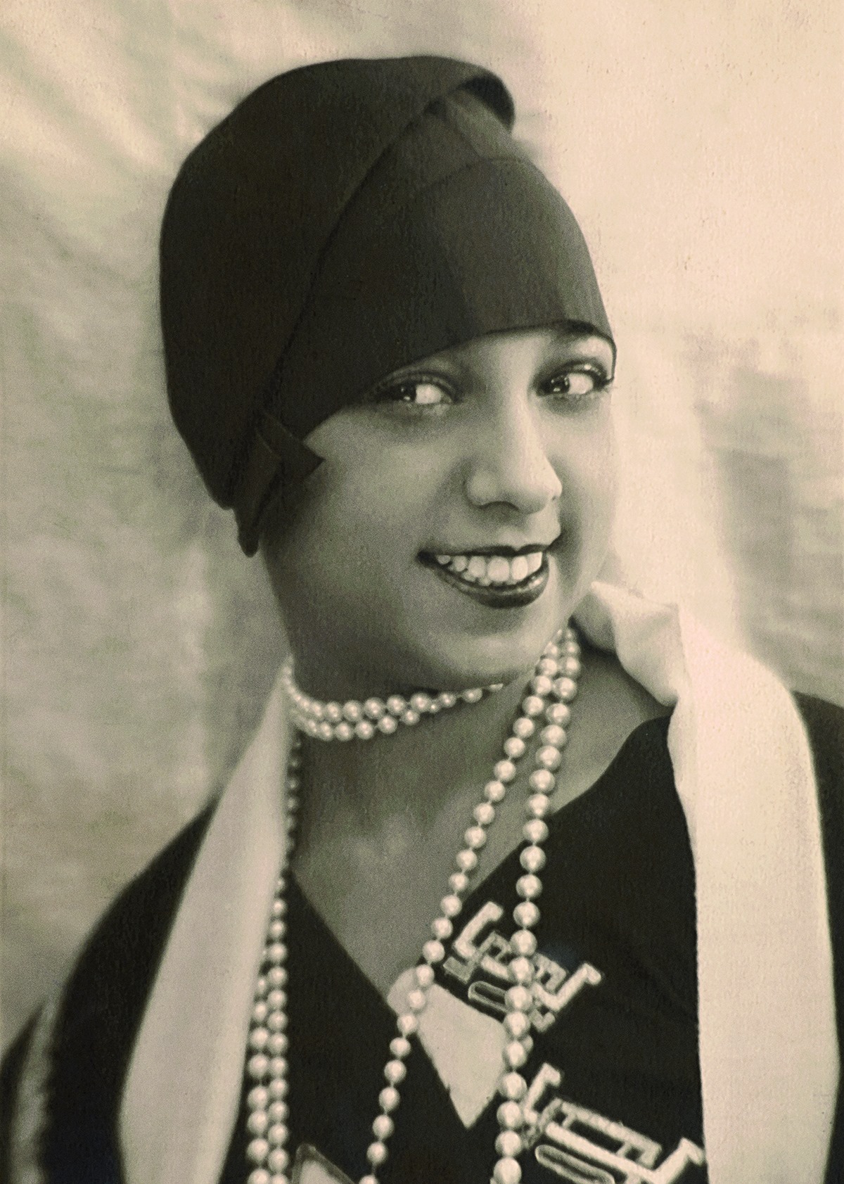 Grayscale portrait photo of Josephine Baker in a black dress with white pearls and a black hat