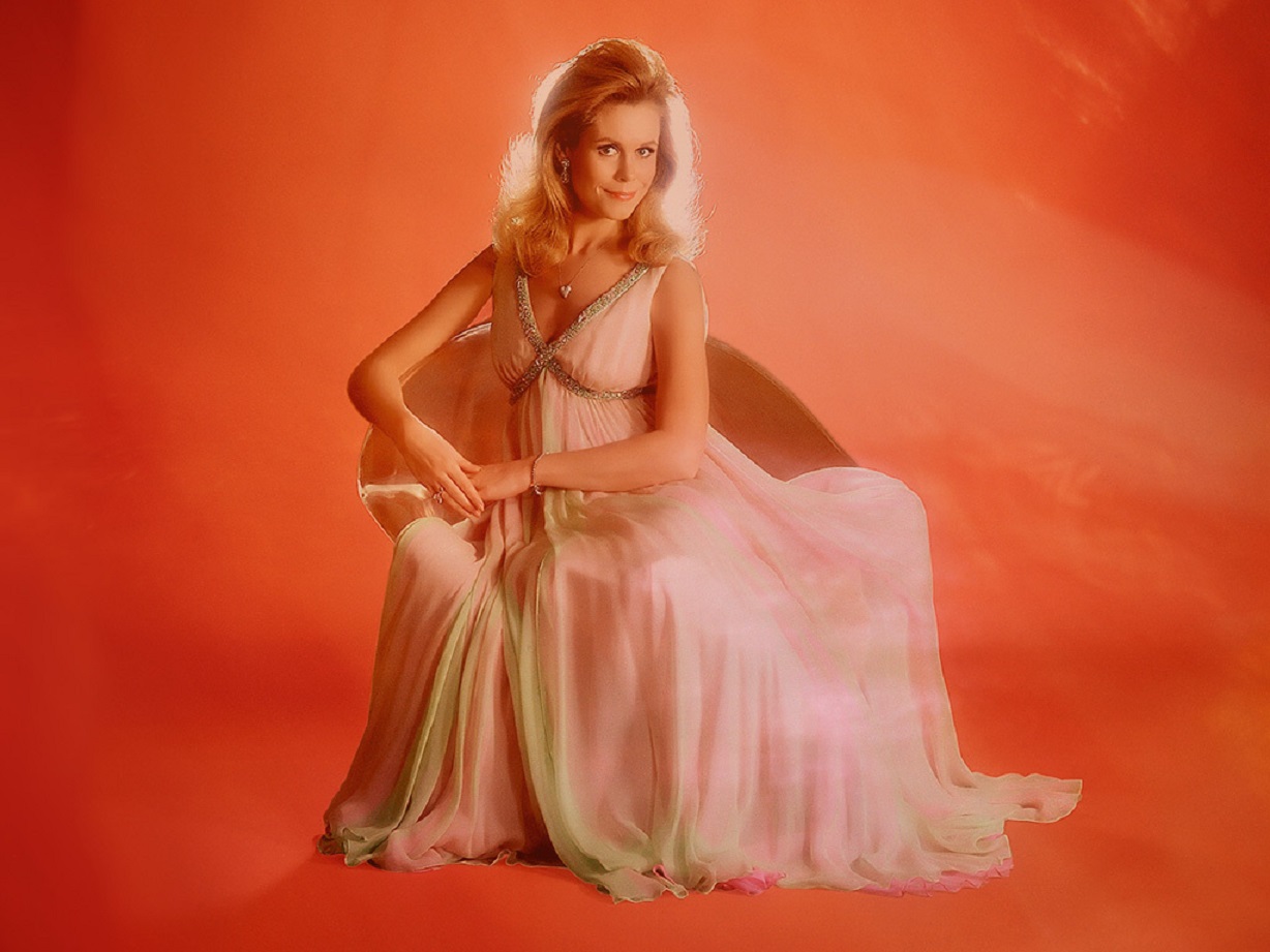 Color portrait of Elizabeth Montgomery wearing pink dress on a red background looking at camera.