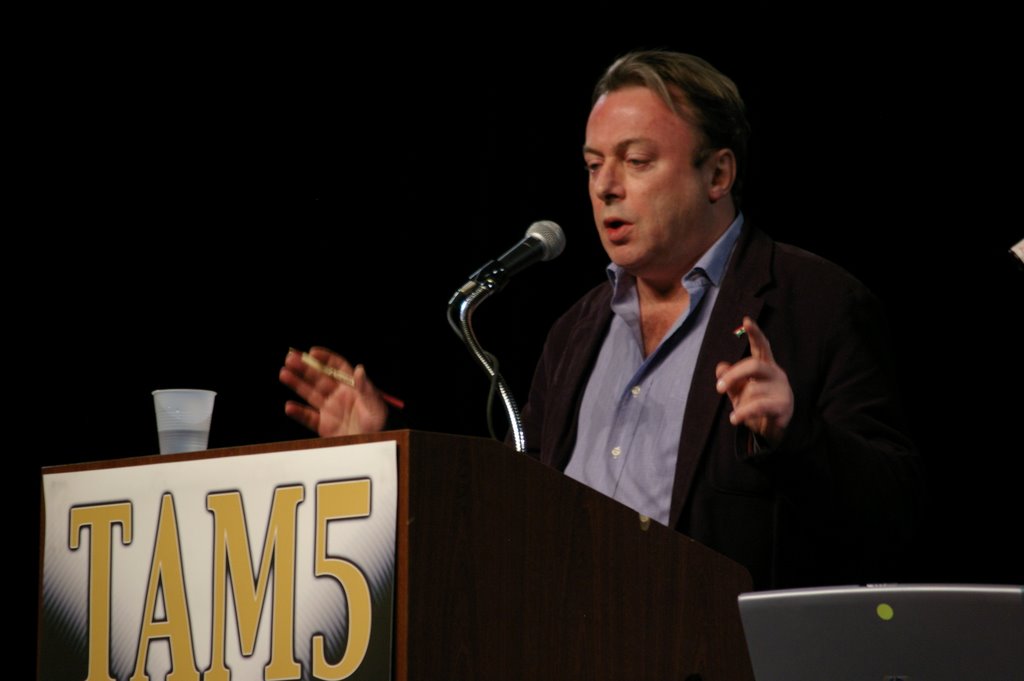 British author and journalist Christopher Hitchens giving a speech wearing a dark jacket blue shirt with his arms raised holding a pen