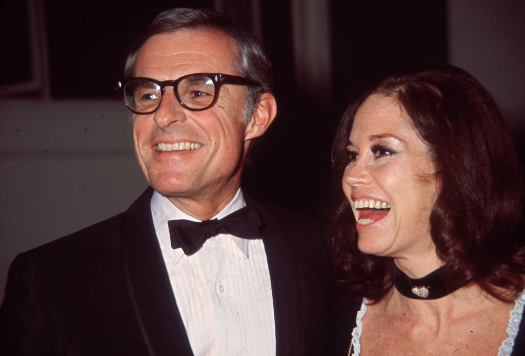 Moore is wearing a low-cut black top with white lace trim, and a black choker, and Tinker wears a tuxedo