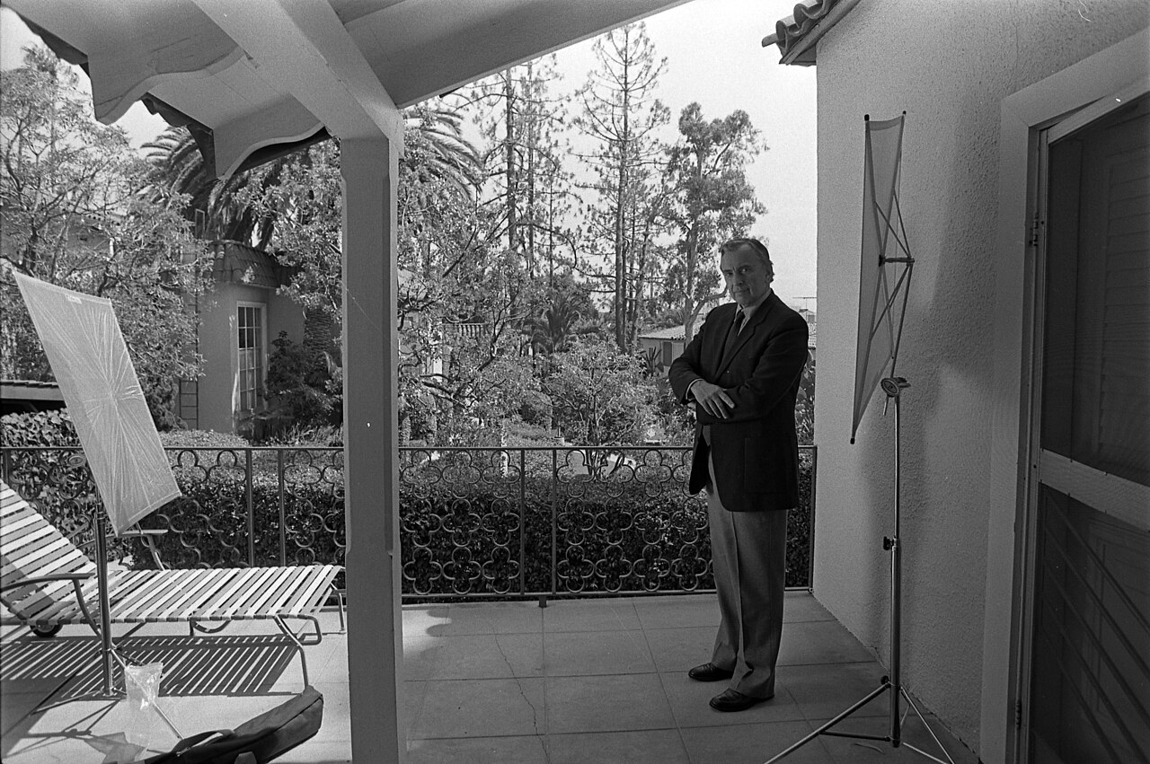 Full-length portrait of Gore Vidal in the backyard of a house standing with his arms crossed wearing a dark suit