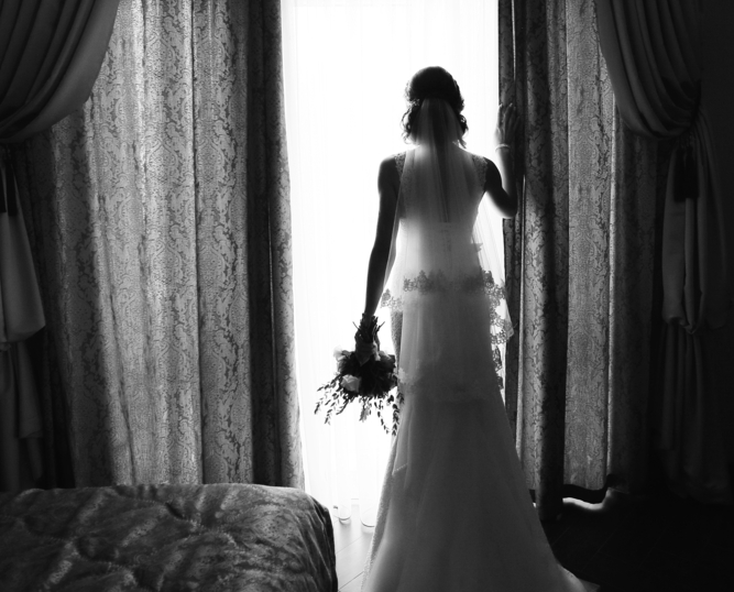 Grayscale photo of a Bride in a wedding gown with her back turned holding a flower. standing by window