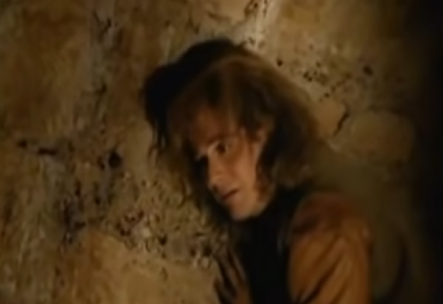 Screenshot: (Mark Umbers) Perkin Warbeck with long hair and brown shirt is standing next to a wall - from Princes in the Tower (2005).
