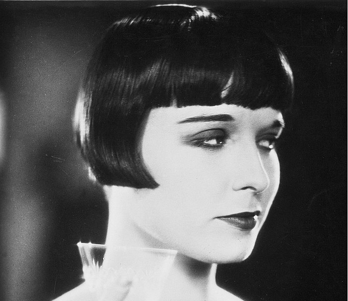 Louise Brooks in a publicity photo for Diary of Lost Girl, holding a glass, wearing black dress, necklace