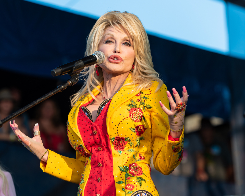 Dolly Parton performs at The Newport Folk Festival in Rhode Island wearing yellow and red dress, microphone in front