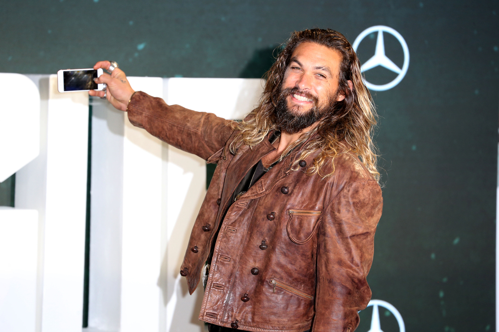 Jason Momoa attends the 'Justice League' photocall at The College in London, England. Wearing brown leathe jacket and holding a mobile.