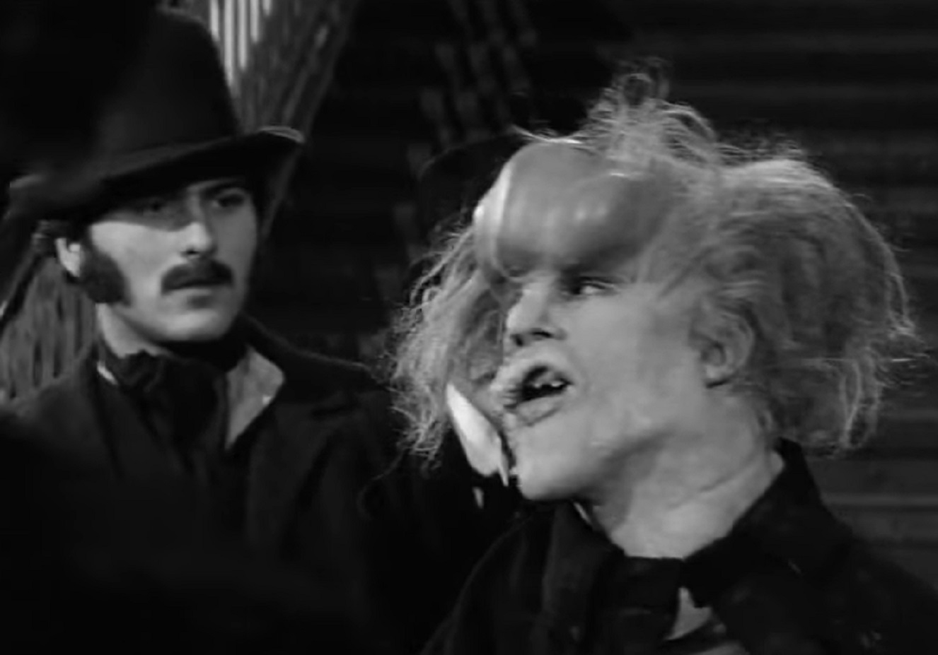 B&W Screenshot of Joseph Merrick (John Hurt) with messy hair is standing next to other man - from Elephant Man (1980)