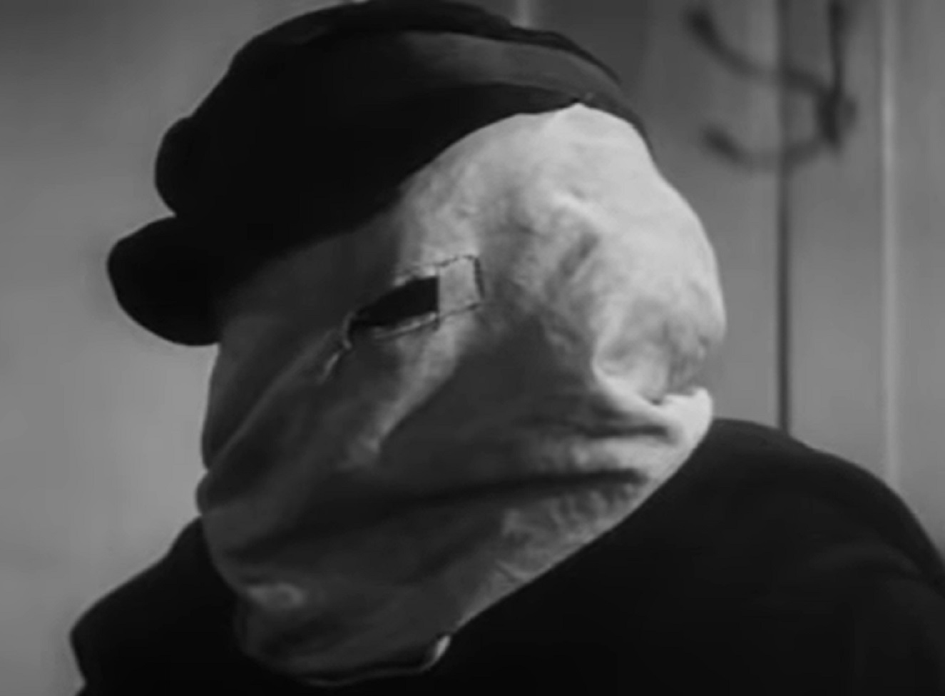 B&W Screenshot of Joseph Merrick (John Hurt) looking at side and wearing hat and white mask over his face - from Elephant Man (1980)