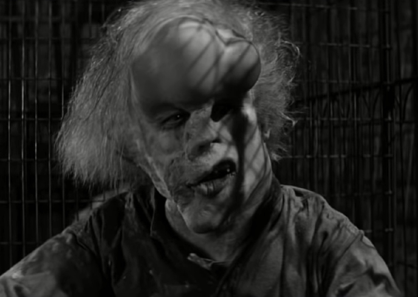 B&W Screenshot of Joseph Merrick (John Hurt) looking at side, seating inside of a cage - from Elephant Man (1980)