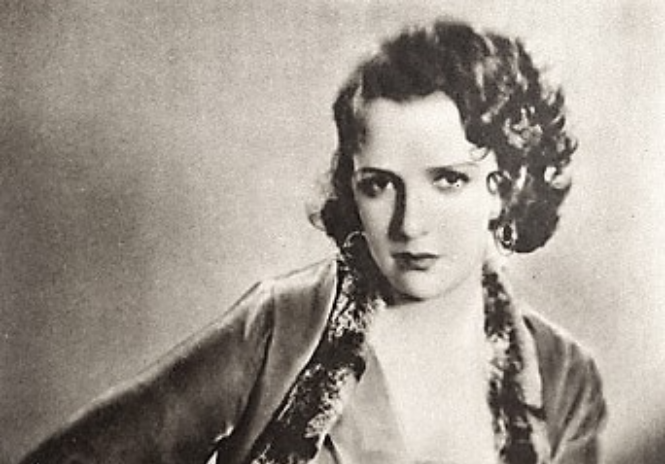 Grayscale portrait photo of the actress Bebe Daniels sitting on a chair wearing a light colored dress and a jacket