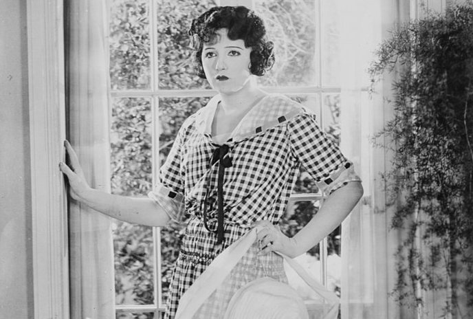 Grayscale Portrait Photo of the Actress Bebe Daniels leaning on a door wearing light colored dress holding a white hat