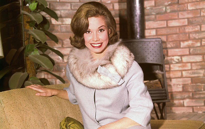 Mary Tyler Moore sitting on a yellow couch wearing a fur collared dress