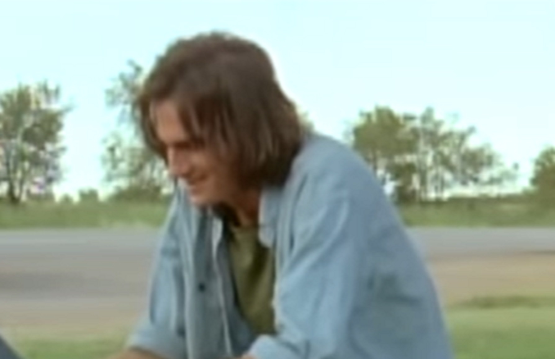Screenshot of James Taylor seating outside with long hair in blue shirt - from Two-Lane Blacktop (1971)