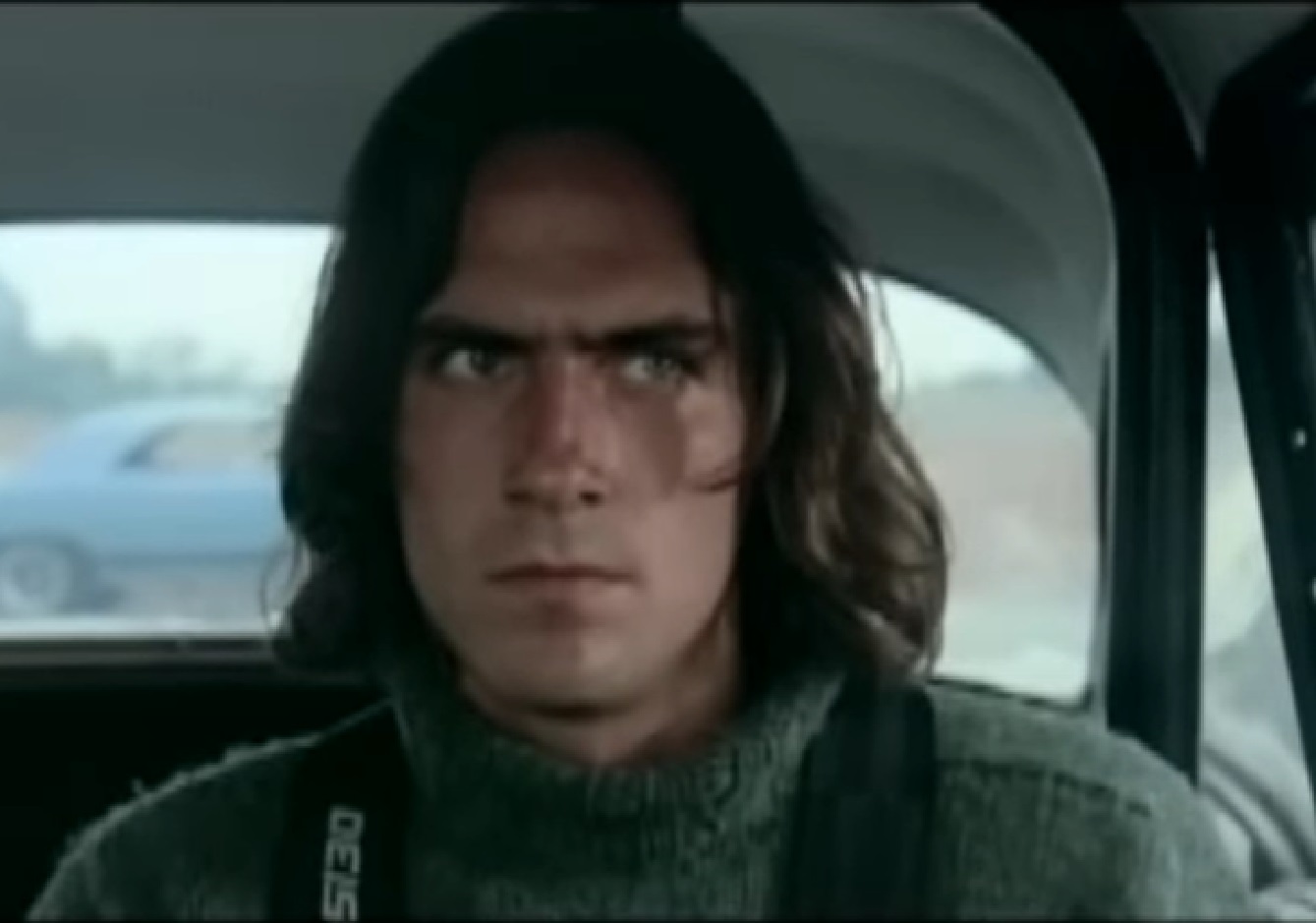 Screenshot of James Taylor with long hair is driving a car and looking upset - from Two-Lane Blacktop (1971)