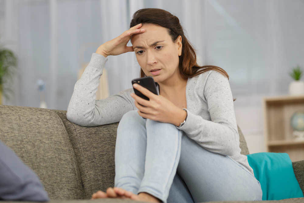 Concerned woman reading message on her smartphone, sitting on a couch