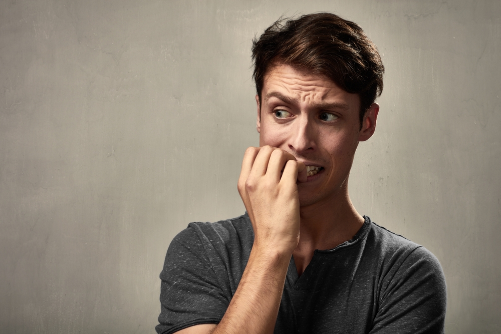 Scared man biting his fingernails, wearing gray t-shirt standing in front of gray background