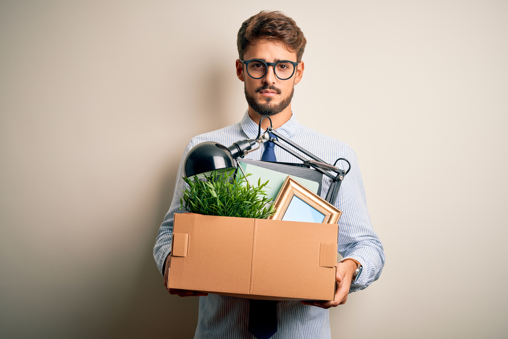 Young businessman with beard fired holding cardboard standing over white background