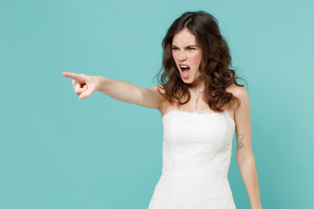 Angry irritated bride in beautiful white wedding dress pointing index finger at front screaming