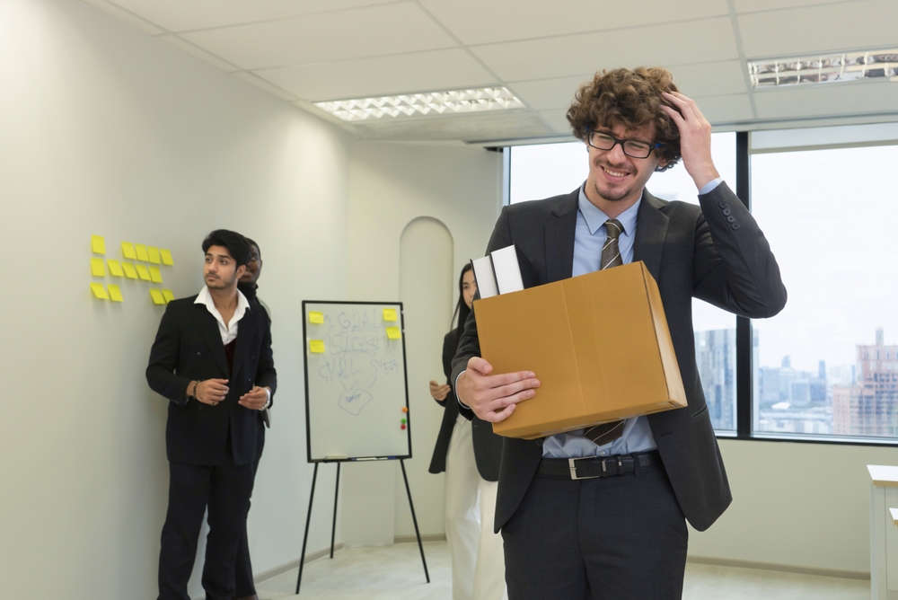 Employee in a suit being fired holding a box of belongings leaving office