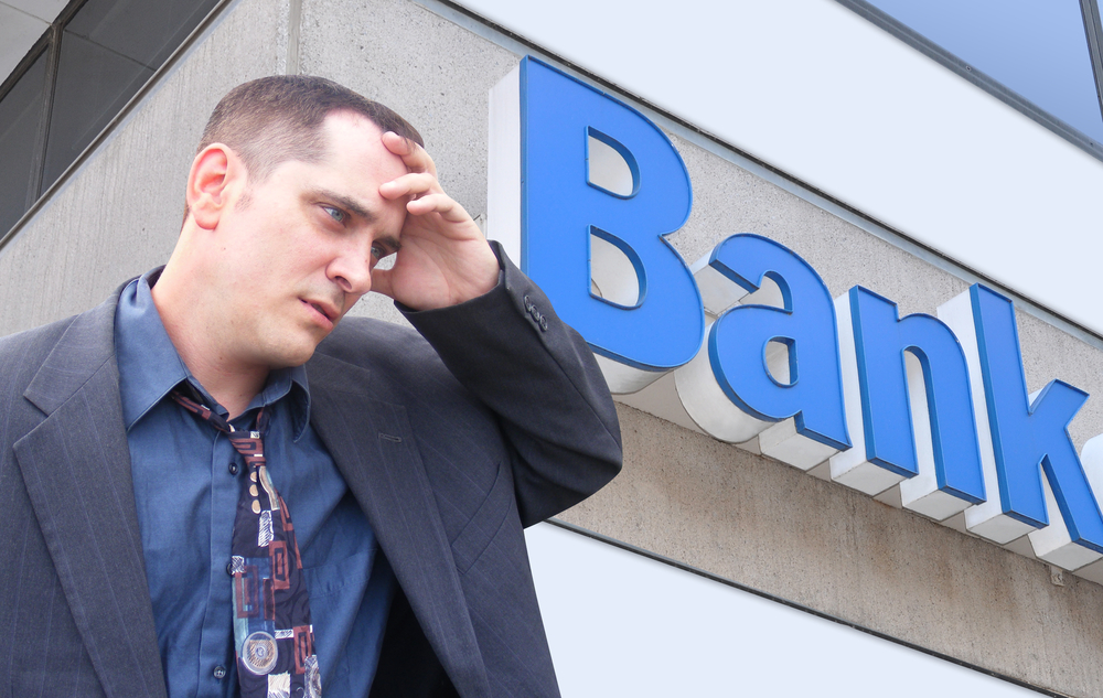 A male employee standing in front of a bank and looking stressed holding his head after being fired from job