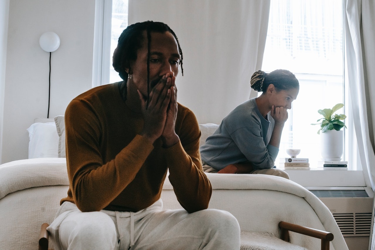 Black man wearing brown sweater seating on the bed with black woman wearing grey sweater looking sad.