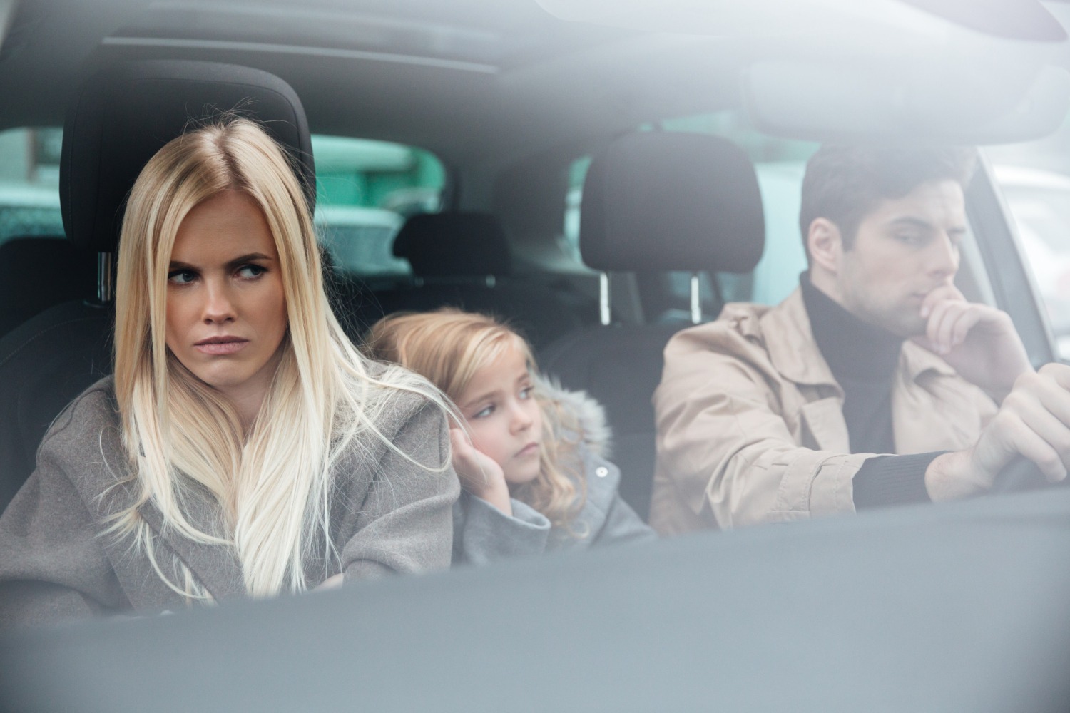 Angry blonde woman seating in the car with man driving with kid on the back seat.