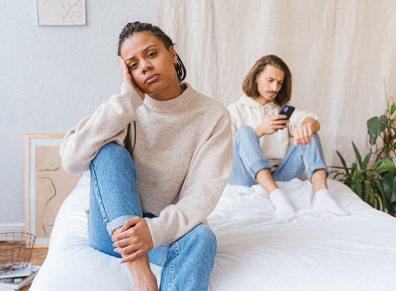 Sad woman seating on the bed with man looking at his phone.
