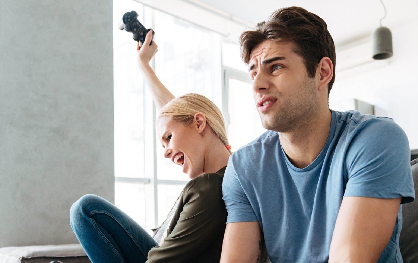 Blonde woman wearing green shirt and jeans looking happy seating next to a upset man wearing blue shirt seating on the sofa.