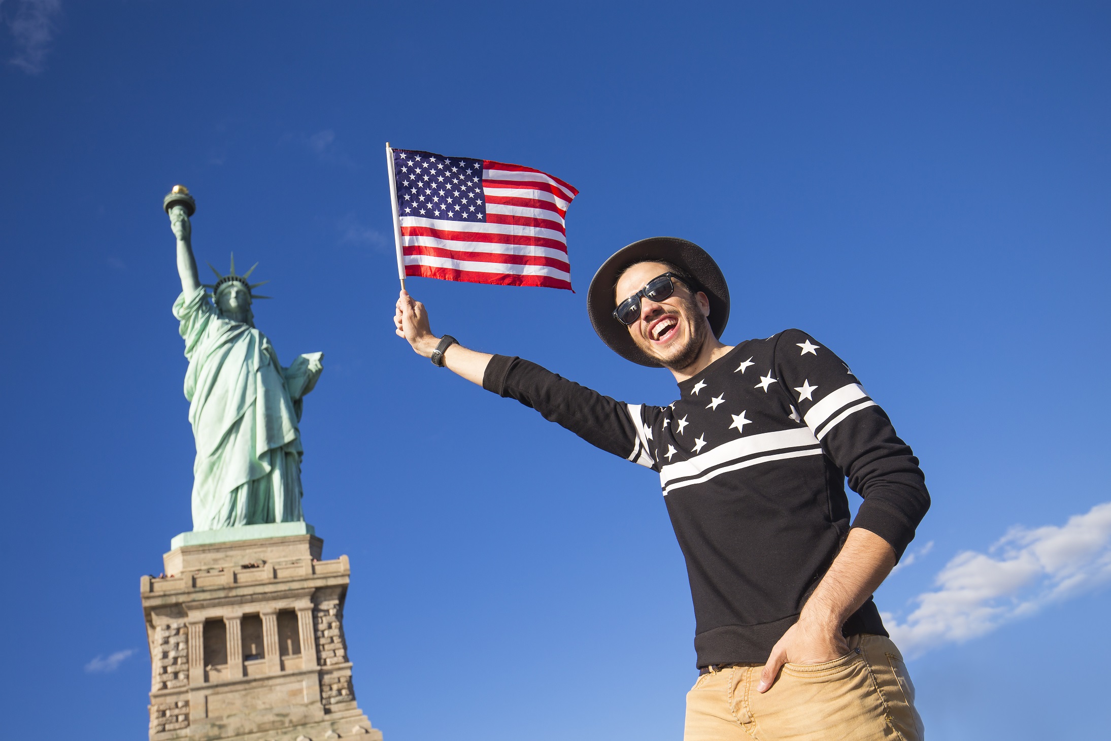 Tourist is holding a small American flag in front of a statue of liberty.