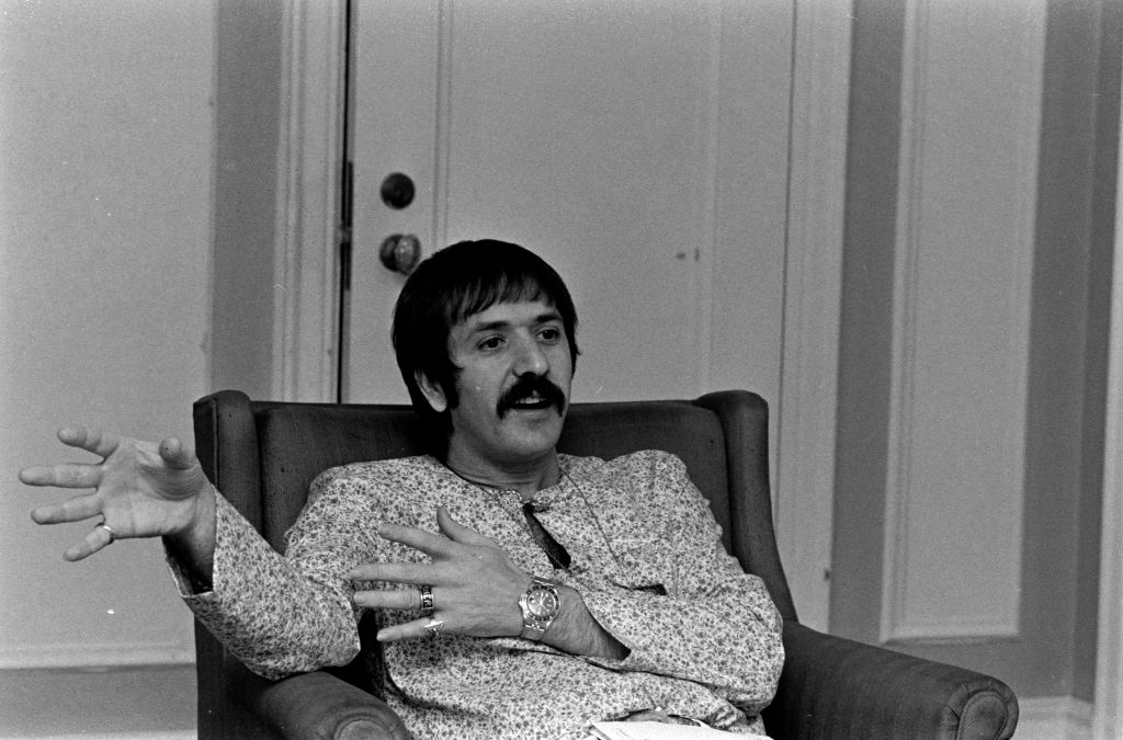 Singer Sonny Bono poses in patterned clothes sitting on an armchair speaking to someone