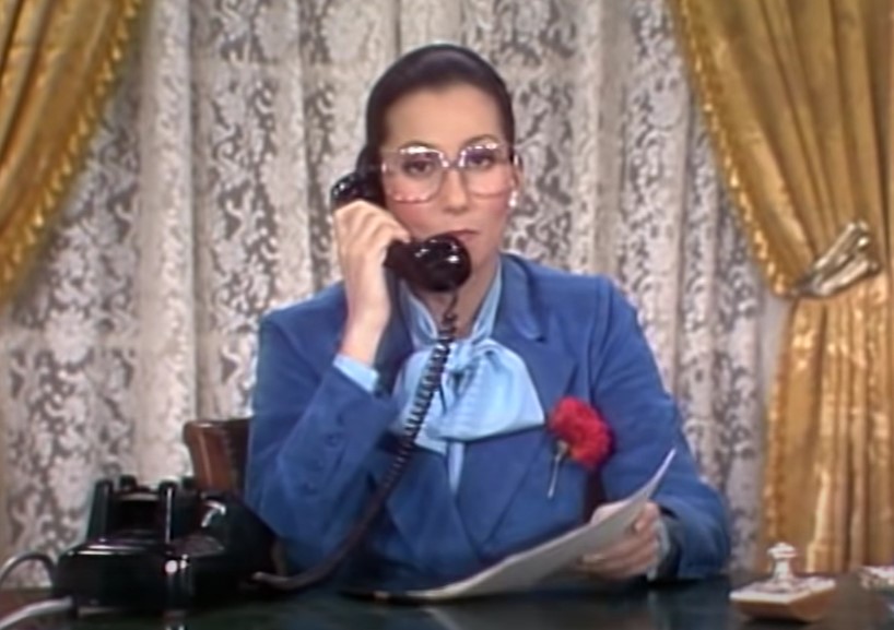 Cher in The Sonny and Cher Comedy Hour sitting on a desk in blue suit and glasses