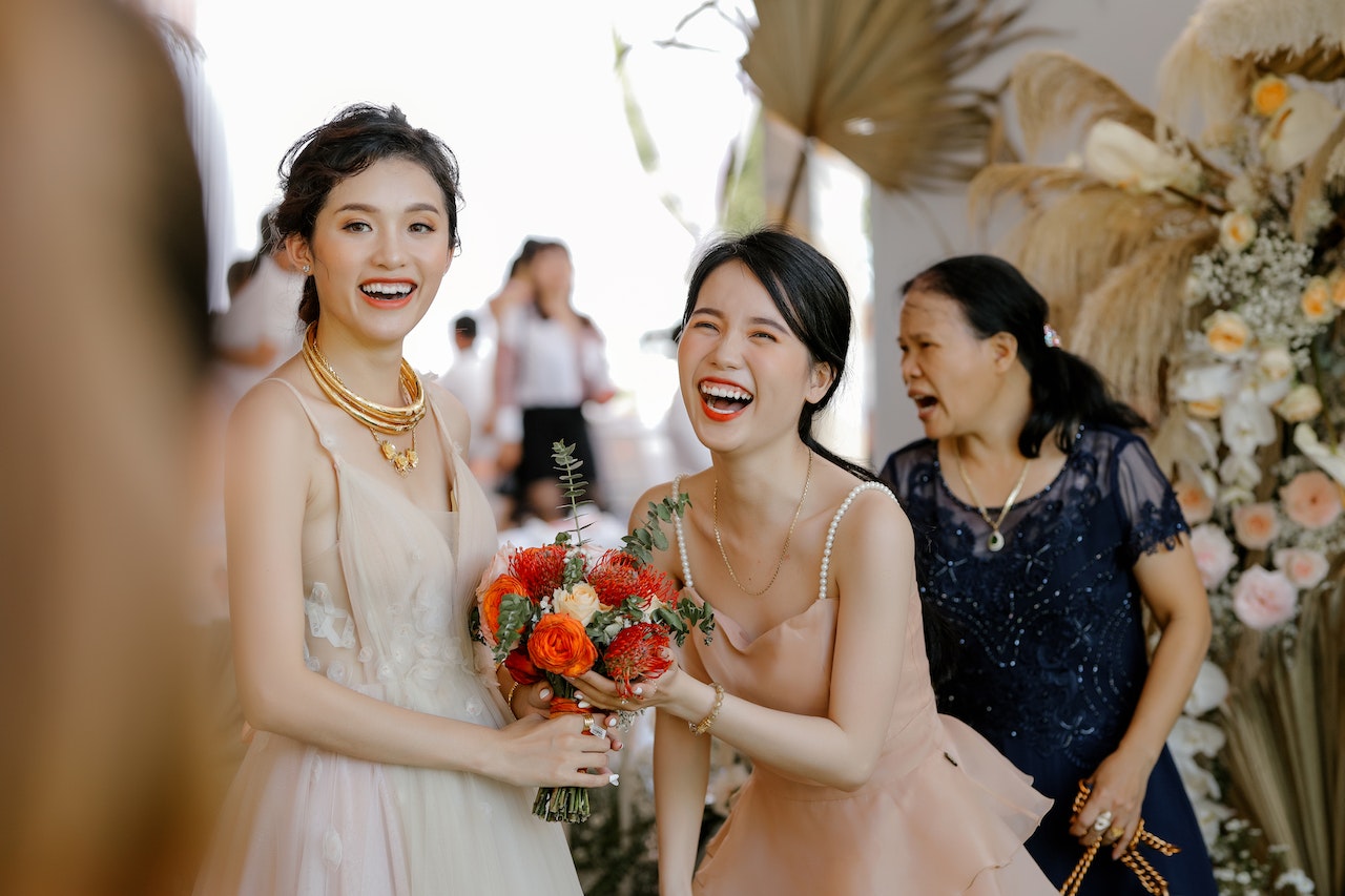 Woman in pink dress is holding a bridal bouquet together with a bride wearing white dress.