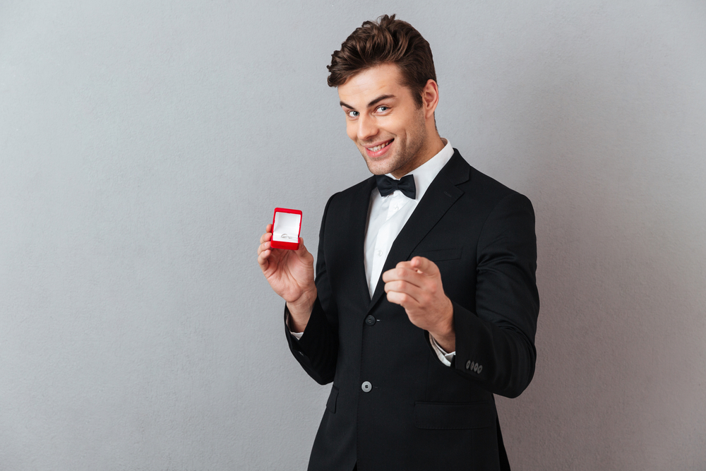 Portrait of a satisfied happy man dressed in tuxedo holding open box with an engagement ring