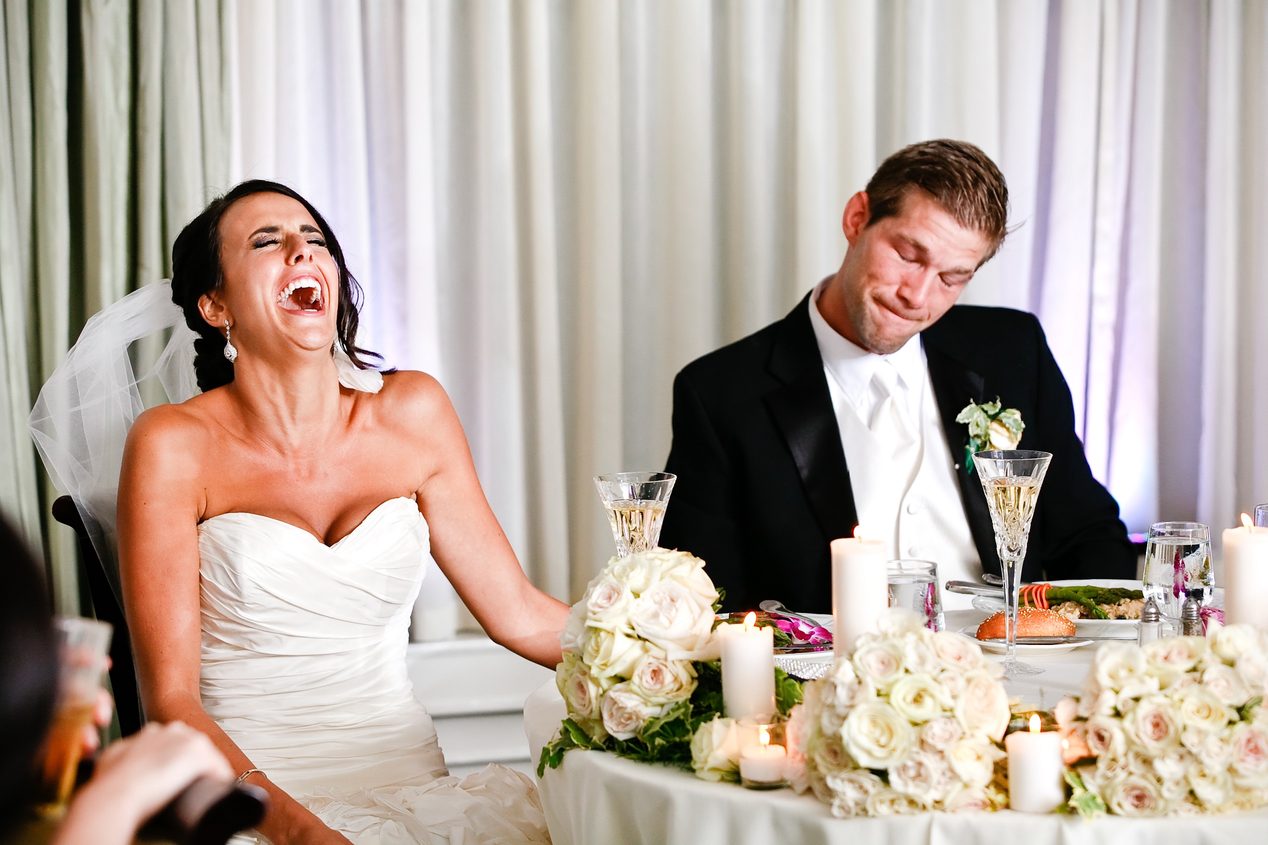 Bride is laughing at wedding reception seating next to groom looking surprised.