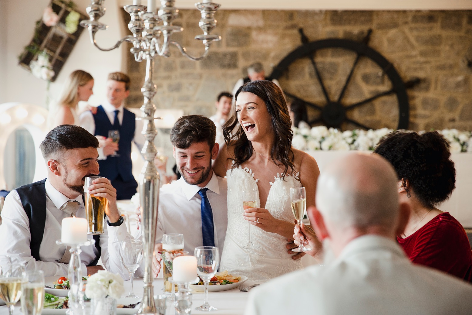 Happy, newly wed couple are seating with guests at their wedding meal and smiling.