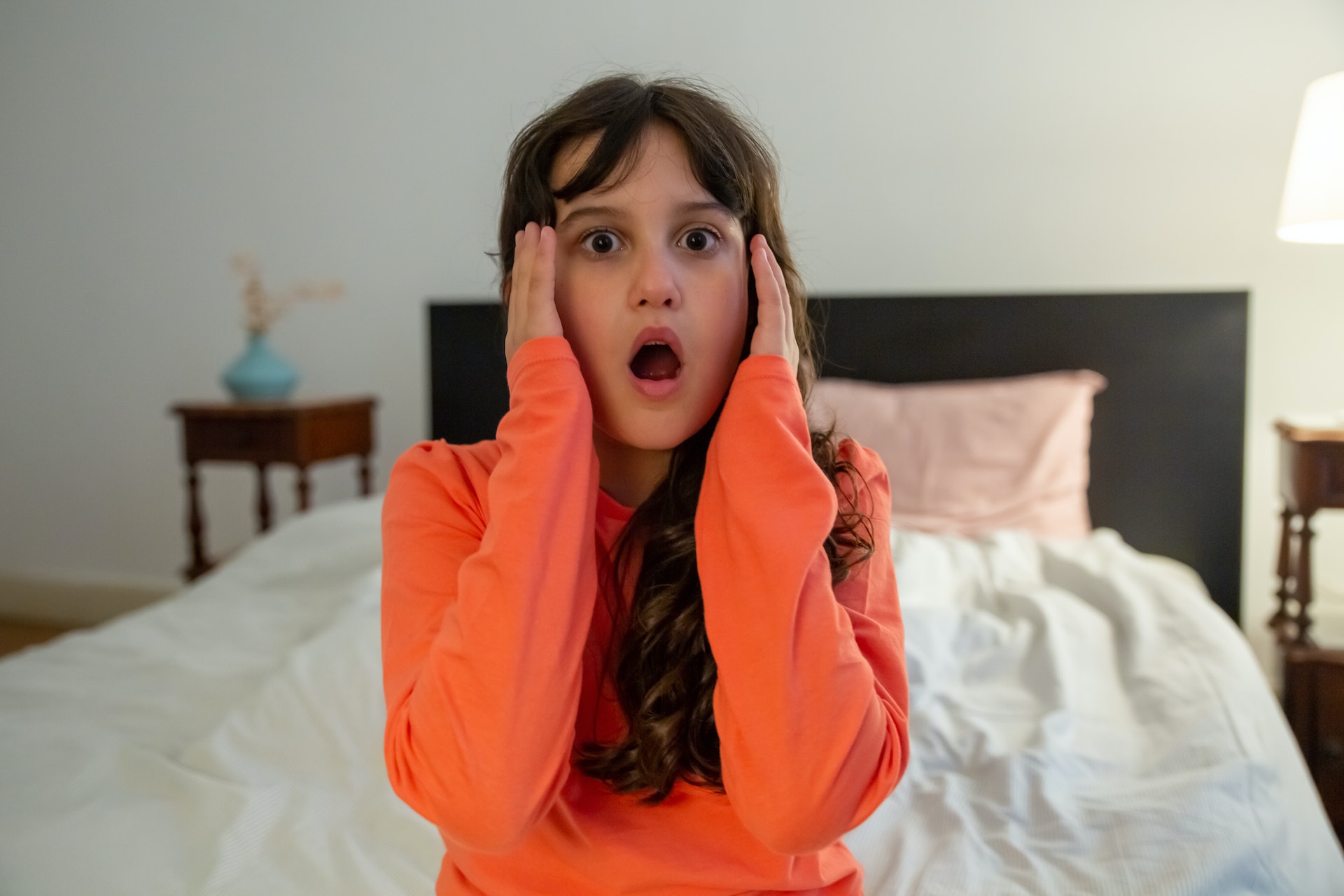 Shocked girl in orange long sleeve shirt sitting on bed