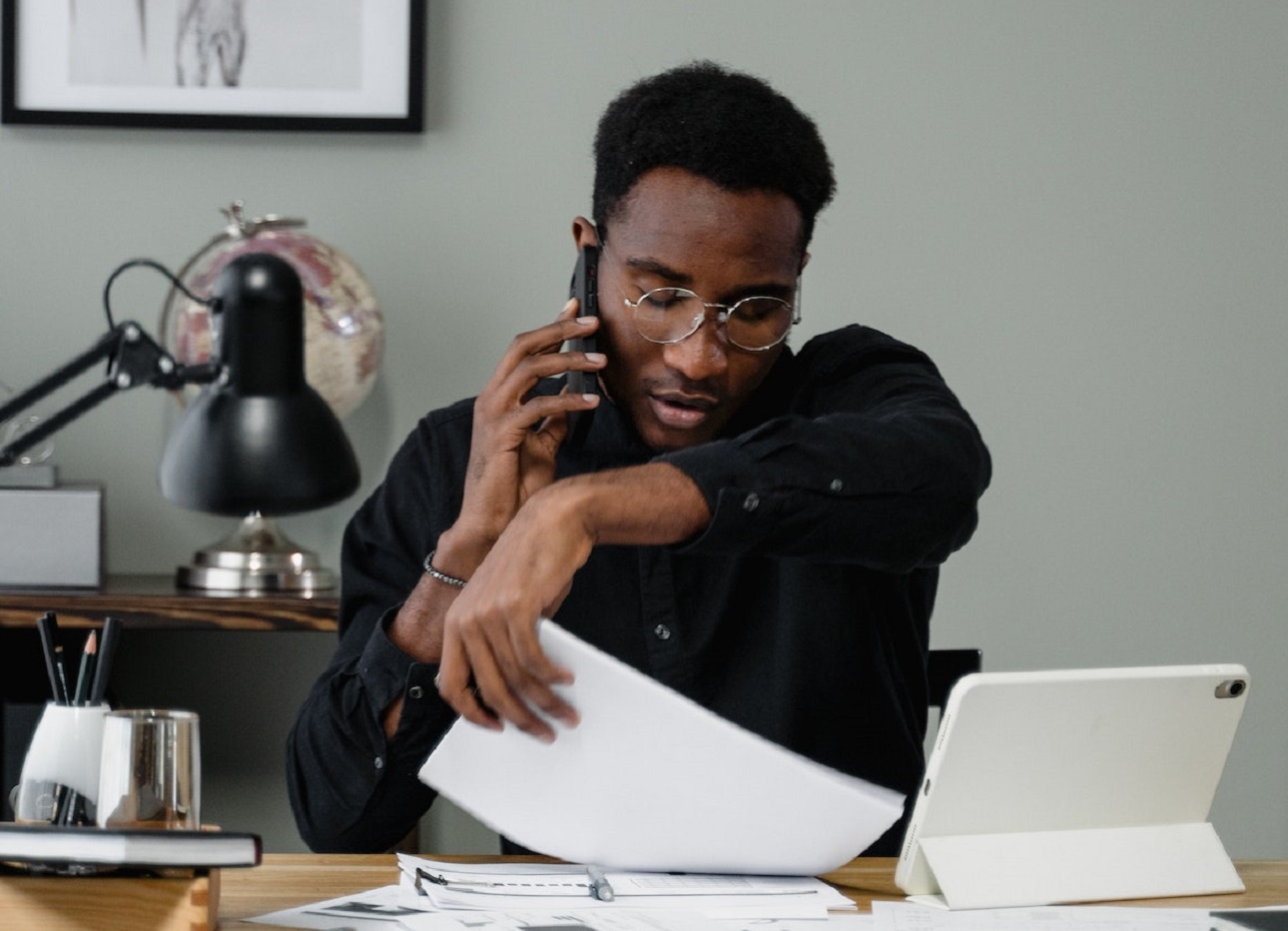 Man is looking at papers at his desk while speaking on cell phone.