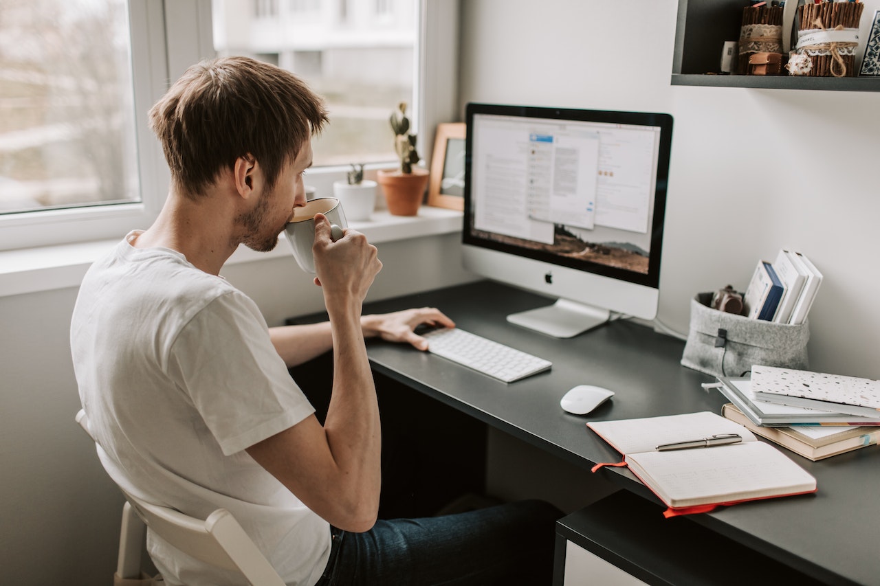 Man is drinking coffee and working on his PC ,seating on the desk.