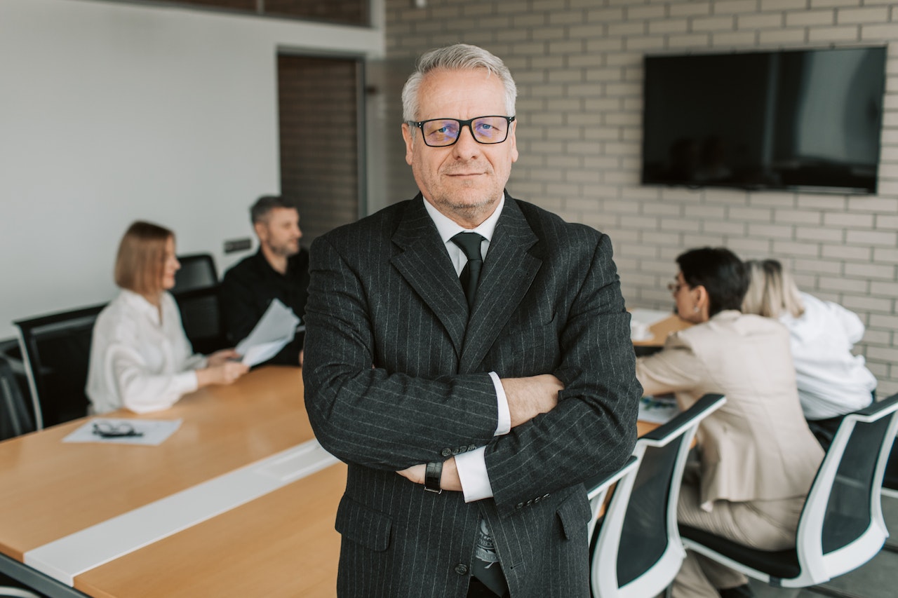 Man in black suit jacket standing in front of a table with people having a meeting.