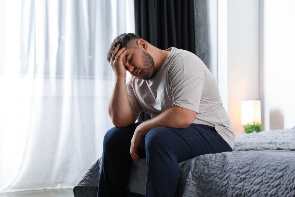Depressed overweight man sitting on bed at home in white t-shirt