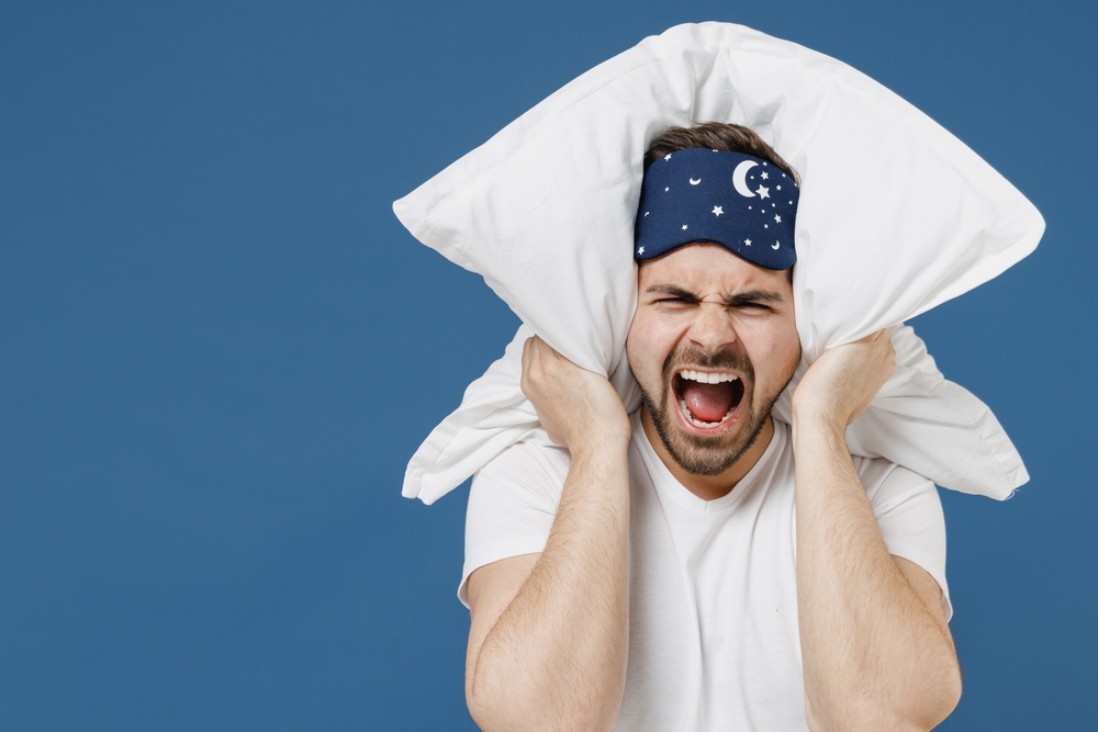 Close up of  young annoyed sad man in pajama   covers his ears with pillow