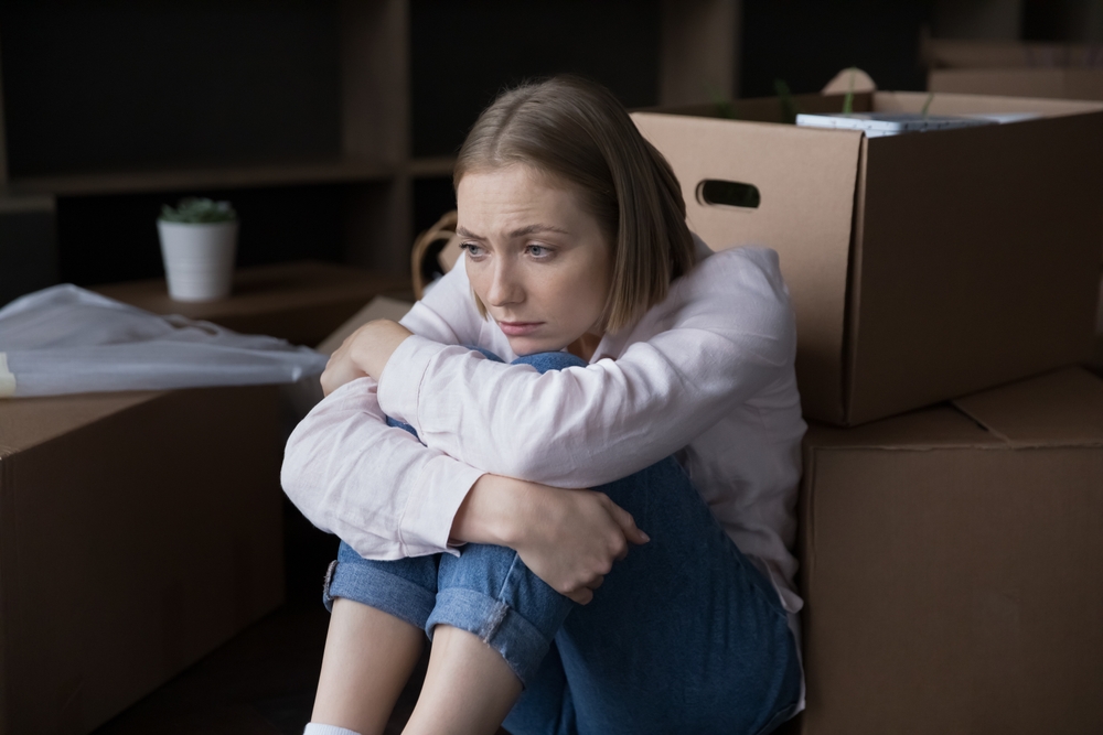 Sad young woman sits near heap of cardboard boxes with personal belongings looks upset goes through divorce