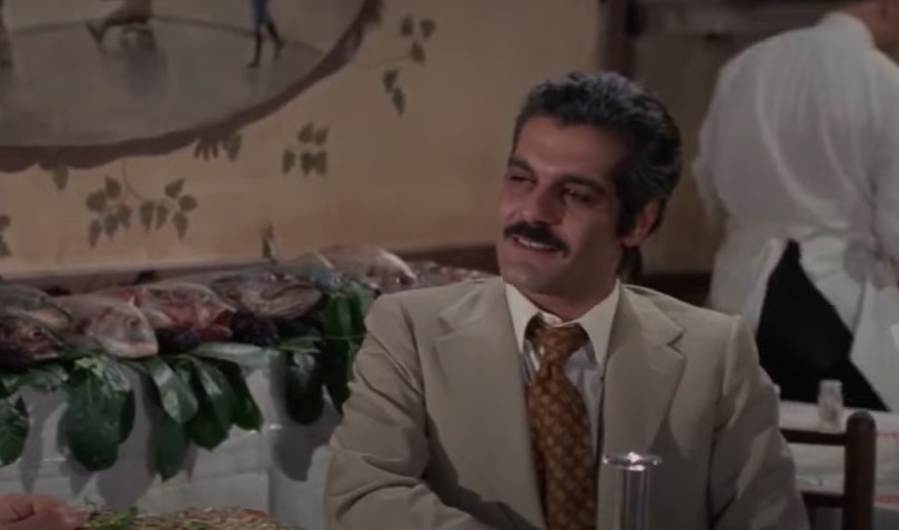 Omar Sharif in The Burglars sitting at a desk in a restaurant speaking to someone