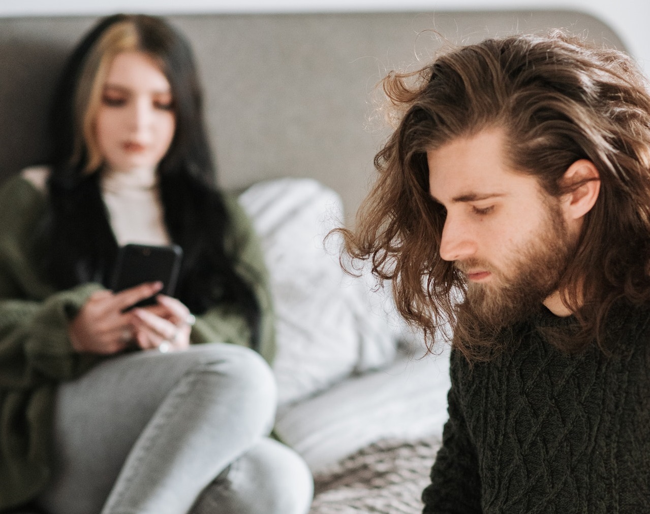 Man in black sweater is seating on the bed near woman in black sweater holding a phone.